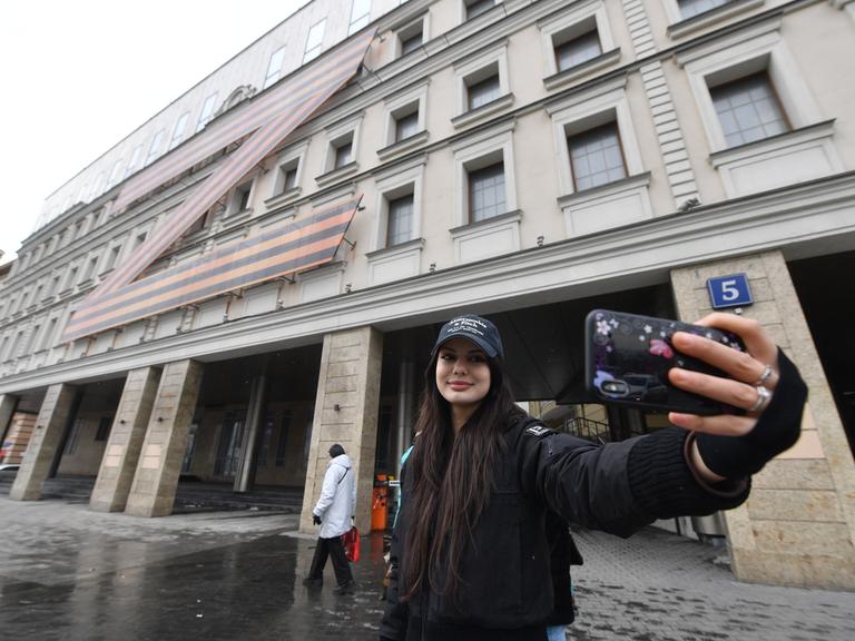 Eine junge Frau fotografiert sich vor einem Gebäude in Moskau, auf dem das Z-Symbol zu sehen ist.