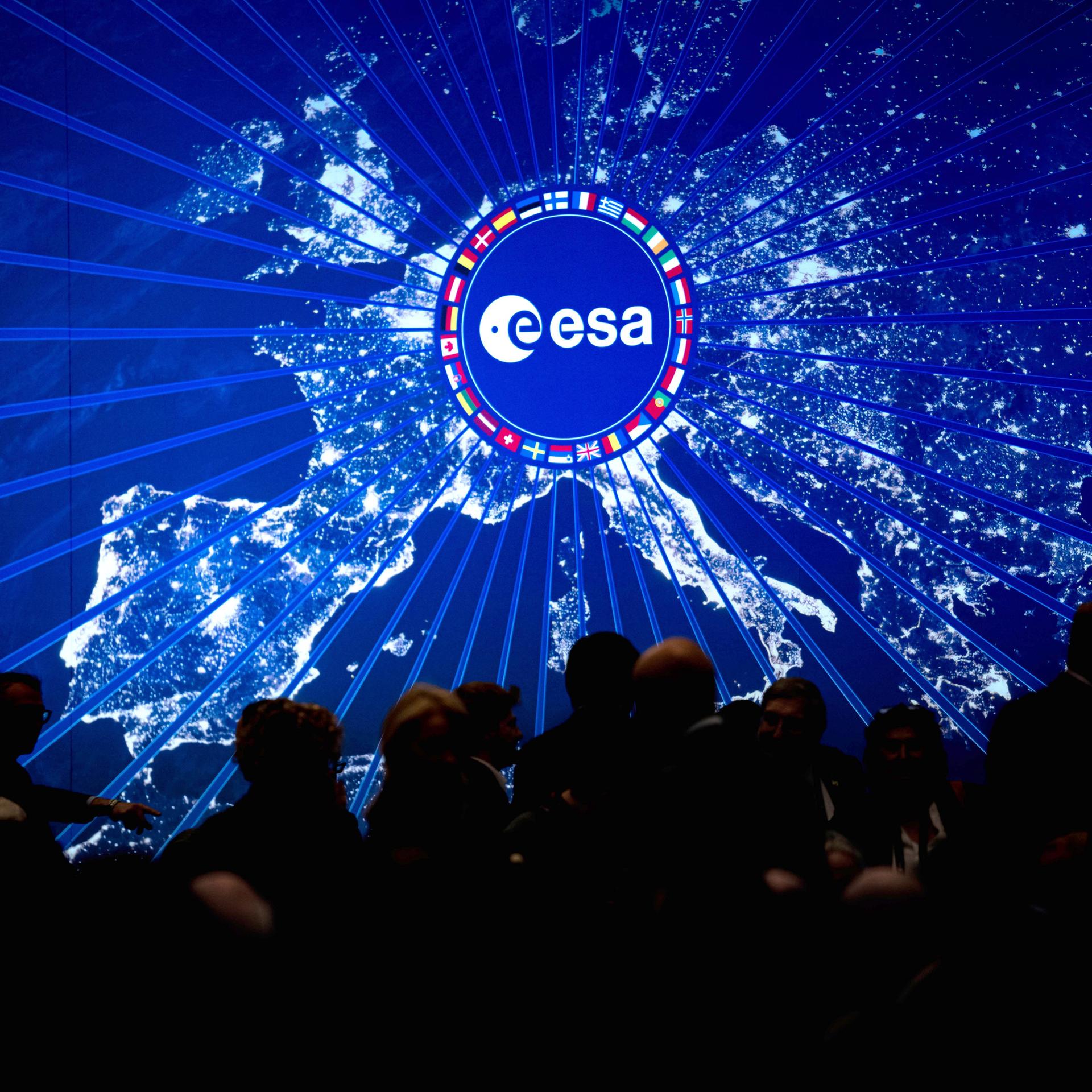 Digitale Darstellung Europas bei Nacht mit hell leuchtenden Städten. In der Mitte befindet sich ein rundes Logo der Europäischen Weltraumorganisation (ESA), umgeben von einem Kreis aus Länderflaggen.