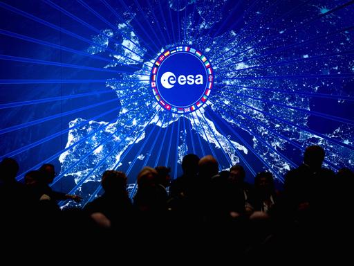 Digitale Darstellung Europas bei Nacht mit hell leuchtenden Städten. In der Mitte befindet sich ein rundes Logo der Europäischen Weltraumorganisation (ESA), umgeben von einem Kreis aus Länderflaggen. Digitale Darstellung Europas bei Nacht mit hell leuchtenden Städten. In der Mitte befindet sich ein rundes Logo der Europäischen Weltraumorganisation (ESA), umgeben von einem Kreis aus Länderflaggen.