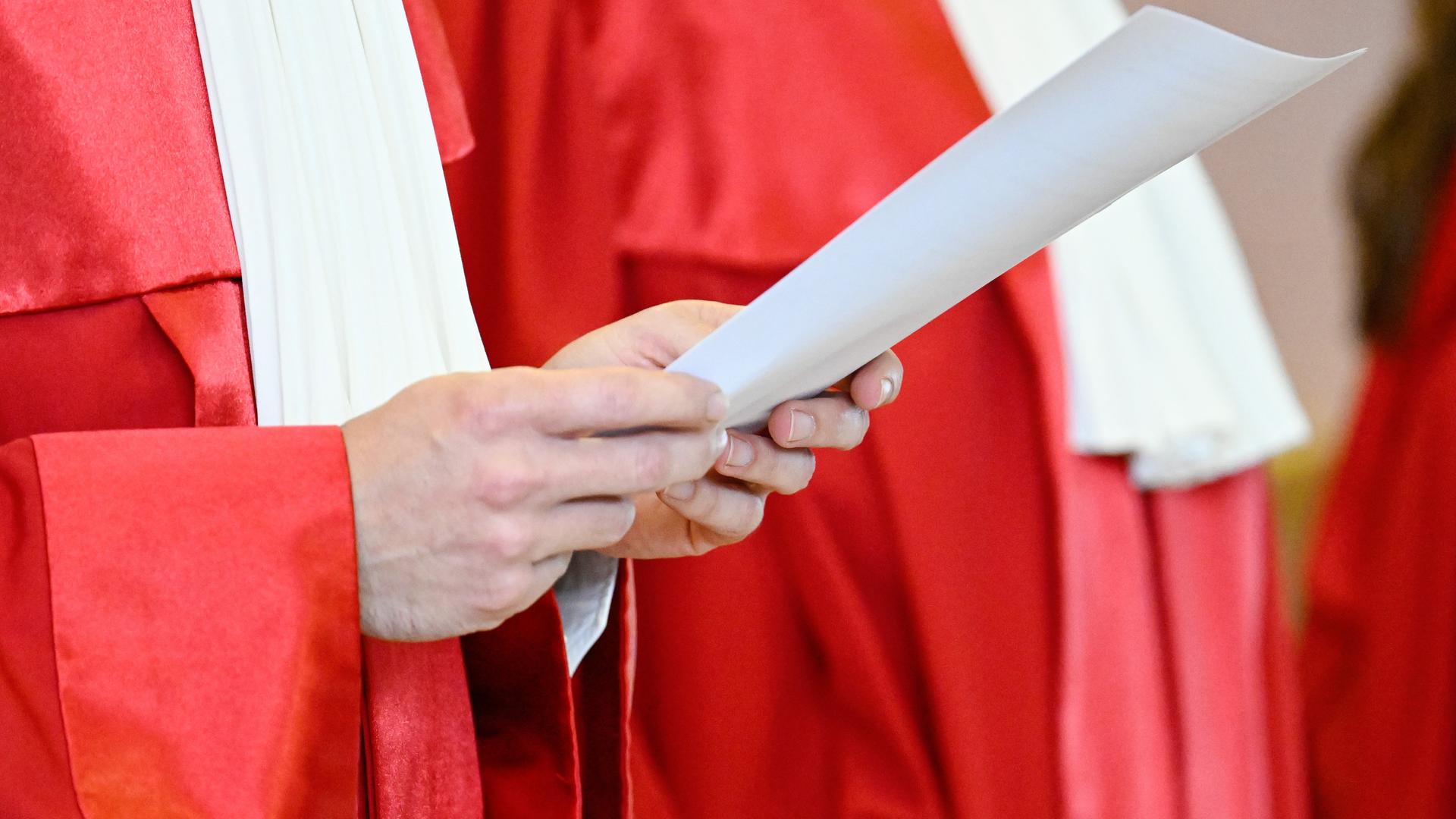 Ein Richter in roter Robe hält ein weißes Blatt bei der Urteilsverkündung in der Hand. Der Bildausschnitt zeigt nur seinen Oberkörper ohne Kopf.