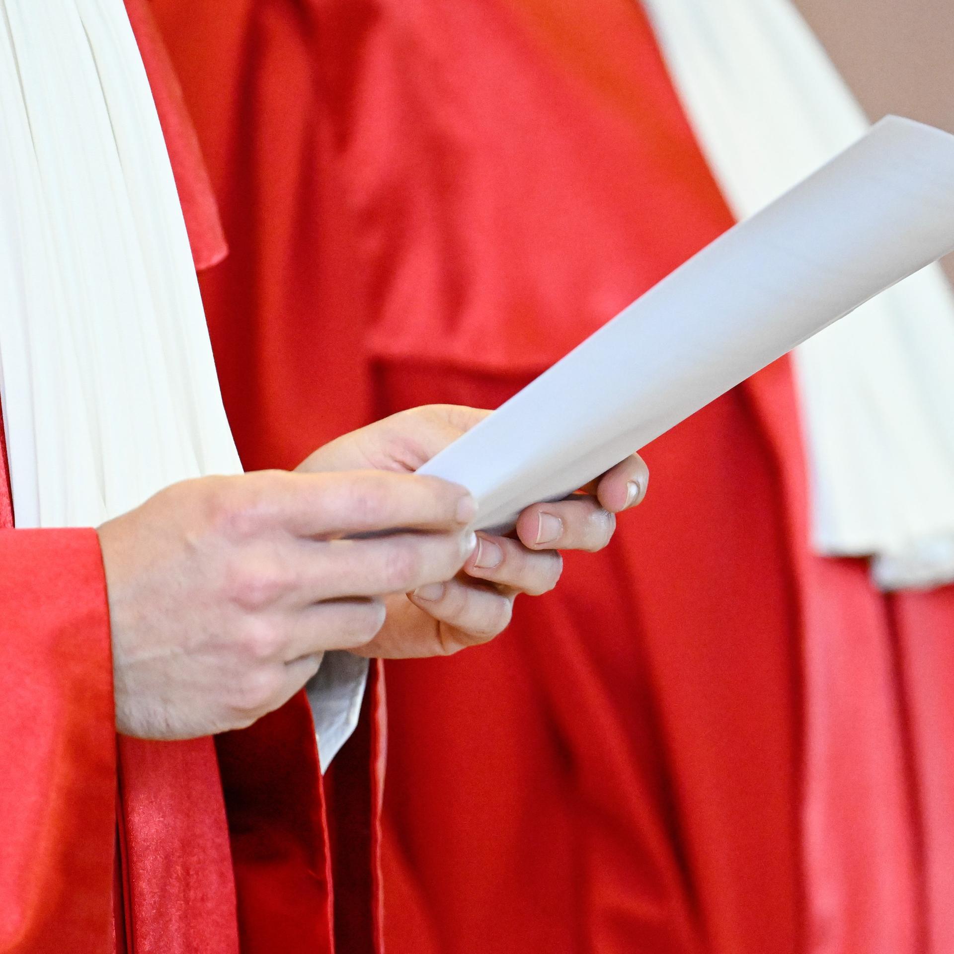 Ein Richter in roter Robe hält ein weißes Blatt bei der Urteilsverkündung in der Hand. Der Bildausschnitt zeigt nur seinen Oberkörper ohne Kopf. Ein Richter in roter Robe hält ein weißes Blatt bei der Urteilsverkündung in der Hand. Der Bildausschnitt zeigt nur seinen Oberkörper ohne Kopf.