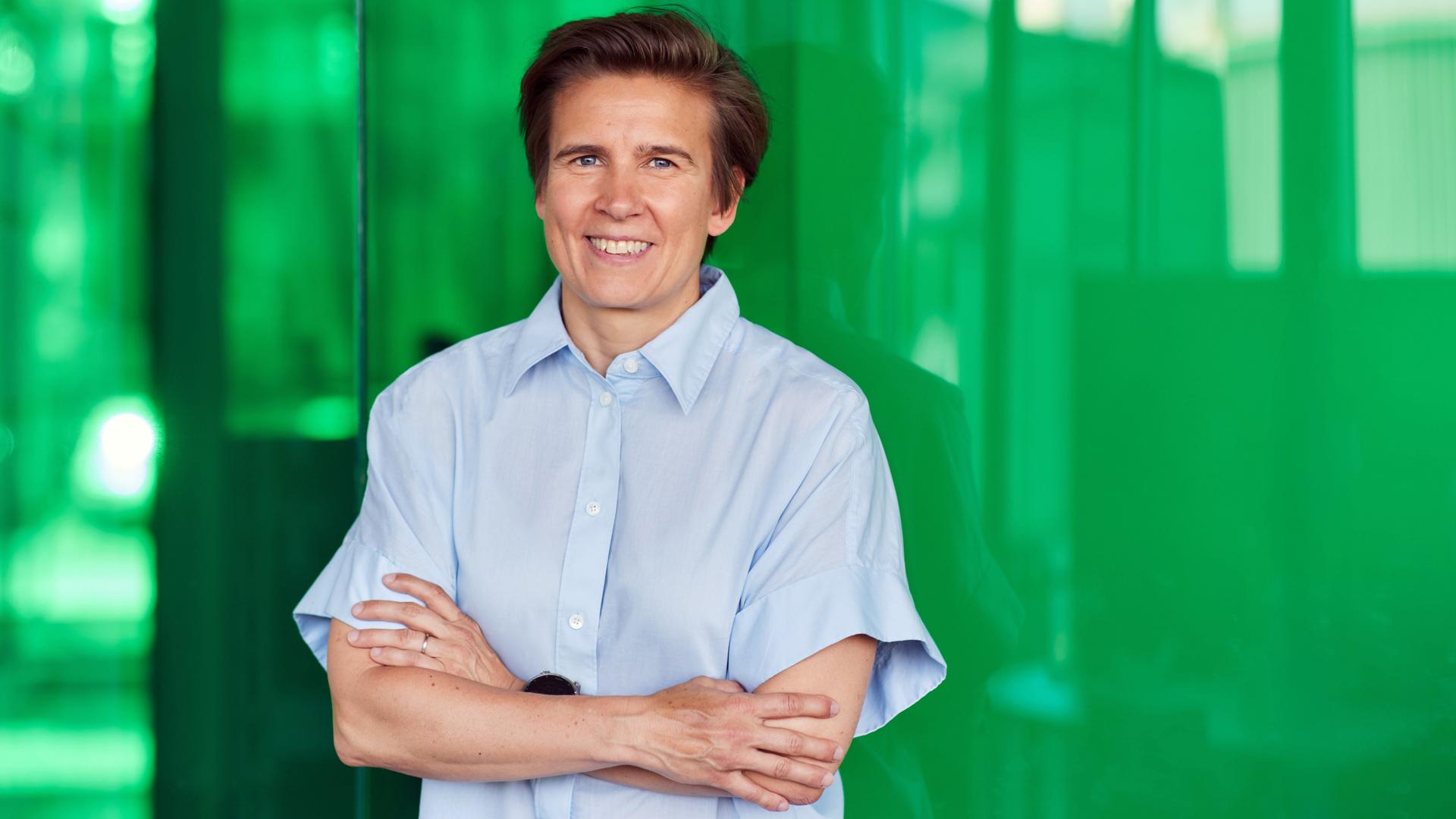 Sara Wickström steht vor einer grünen Wand und hat die Arme vor dem Körper verschränkt.