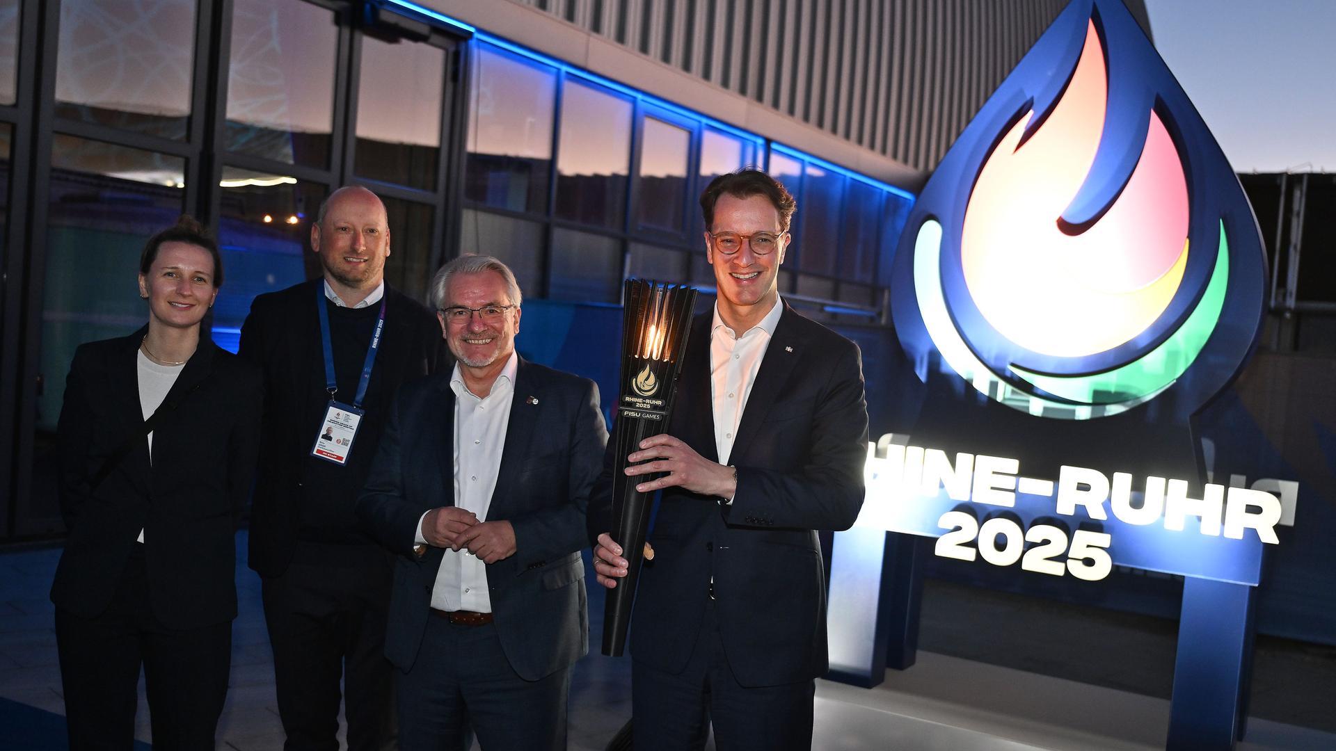 Das Rhein-Ruhr-Organisationsteam der World University Games 2025 um Geschäftsführer Niklas Börger (2.v.l.) posiert mit Hendrik Wüst, Ministerpräsident von NRW, mit der Fackel vor dem Veranstaltungslogo.