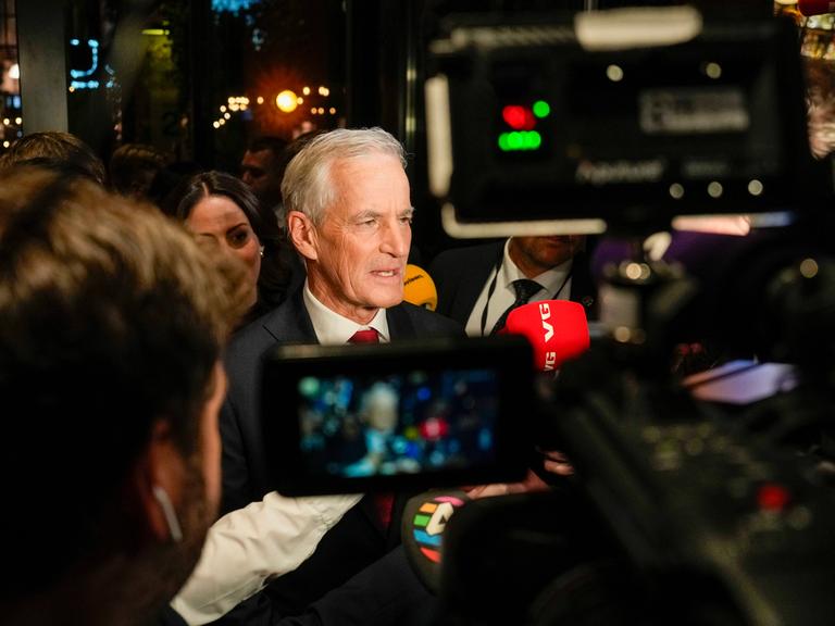 Ein stattlicher Mann im Anzug mit weiß-grauen Haaren steht zwischen vielen Kameras im Gedränge. Weitere gutgekleidete Menschen umgeben ihn. Ein stattlicher Mann im Anzug mit weiß-grauen Haaren steht zwischen vielen Kameras im Gedränge. Weitere gutgekleidete Menschen umgeben ihn.