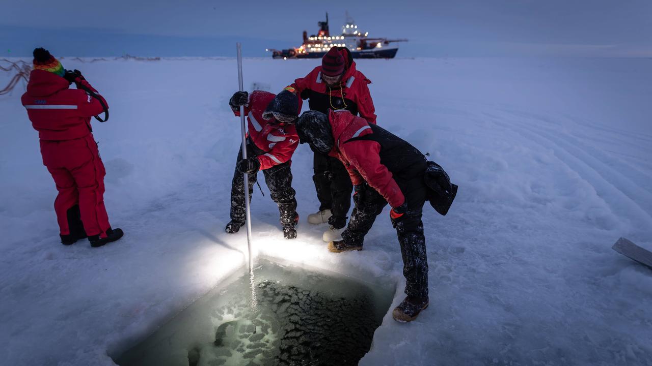Wissenschaftler der Mosaic-Expedition nehmen Proben an einem rechteckigen Loch im Eis; im Hintergrund die "Polarstern" 
