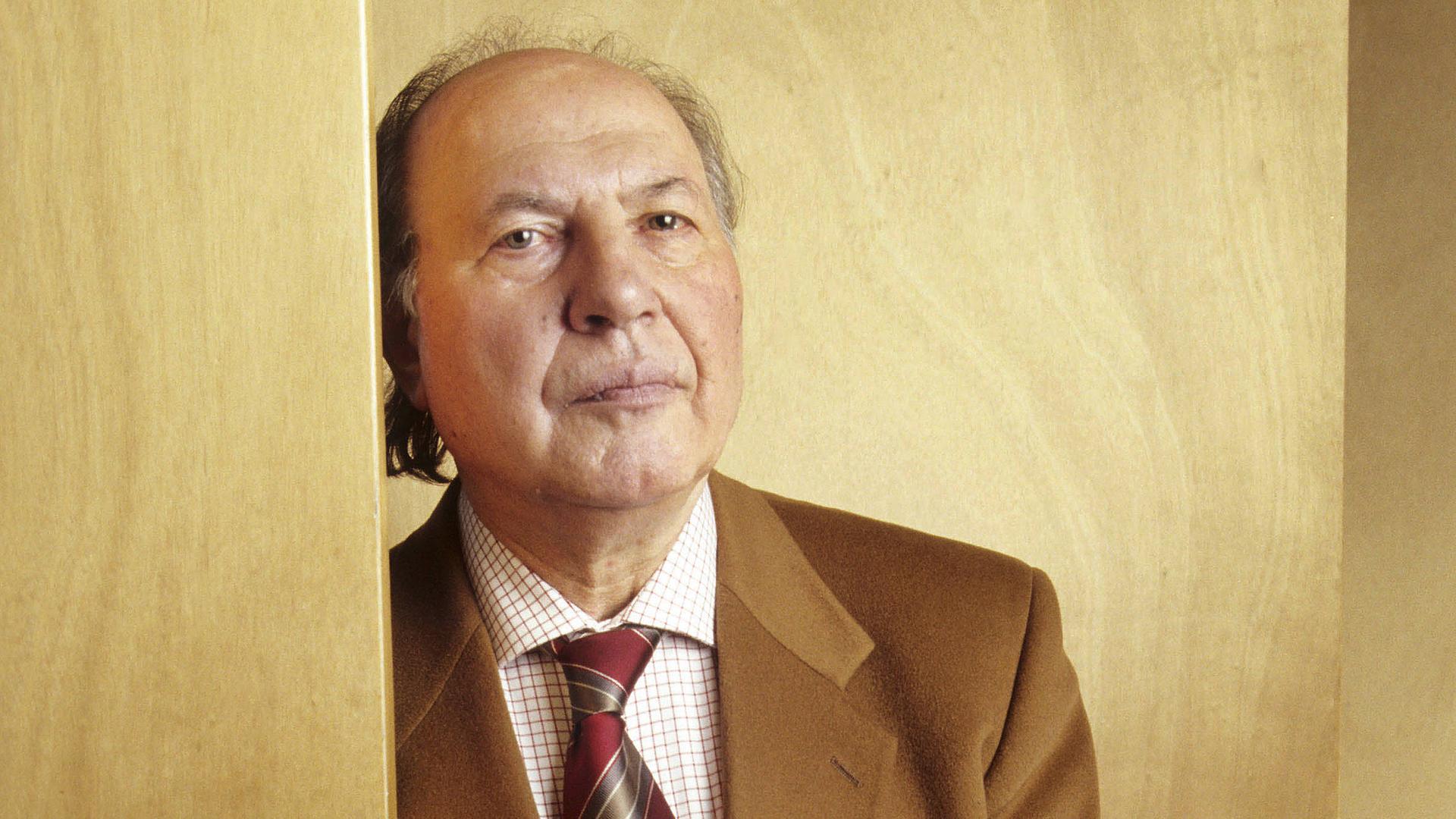 Imre Kertesz lehnt an einer holzvertäfelten Wand und blickt ernst in die Kamera