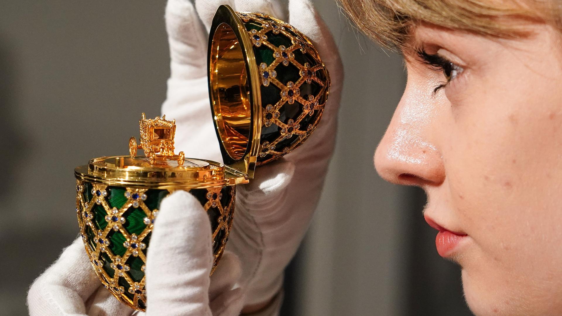 Ein geöffnetes vergoldetes und mit Swarovski-Steinen besetztes Requisiten-Ei aus dem James-Bond-Film "Octopussy" von 1983 wird von einer jungen Frau mit weißen Handschuhen hochgehalten. 