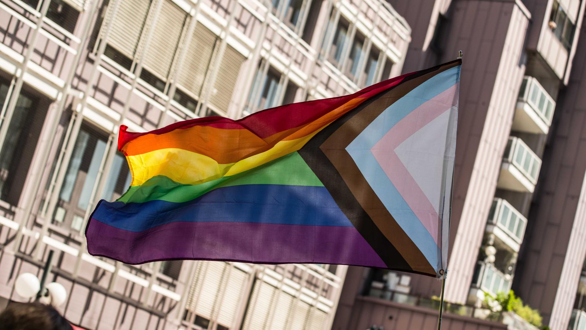 Eine Trans-Pride-Fahne weht im Wind, aufgenommen während einer CSD-Parade in München. Die traditionelle Regenbogenfahne ist ergänzt um einen dreieckigen Einschub, die jeweiligen Farben symbolisieren Teile des queeren Spektrums.