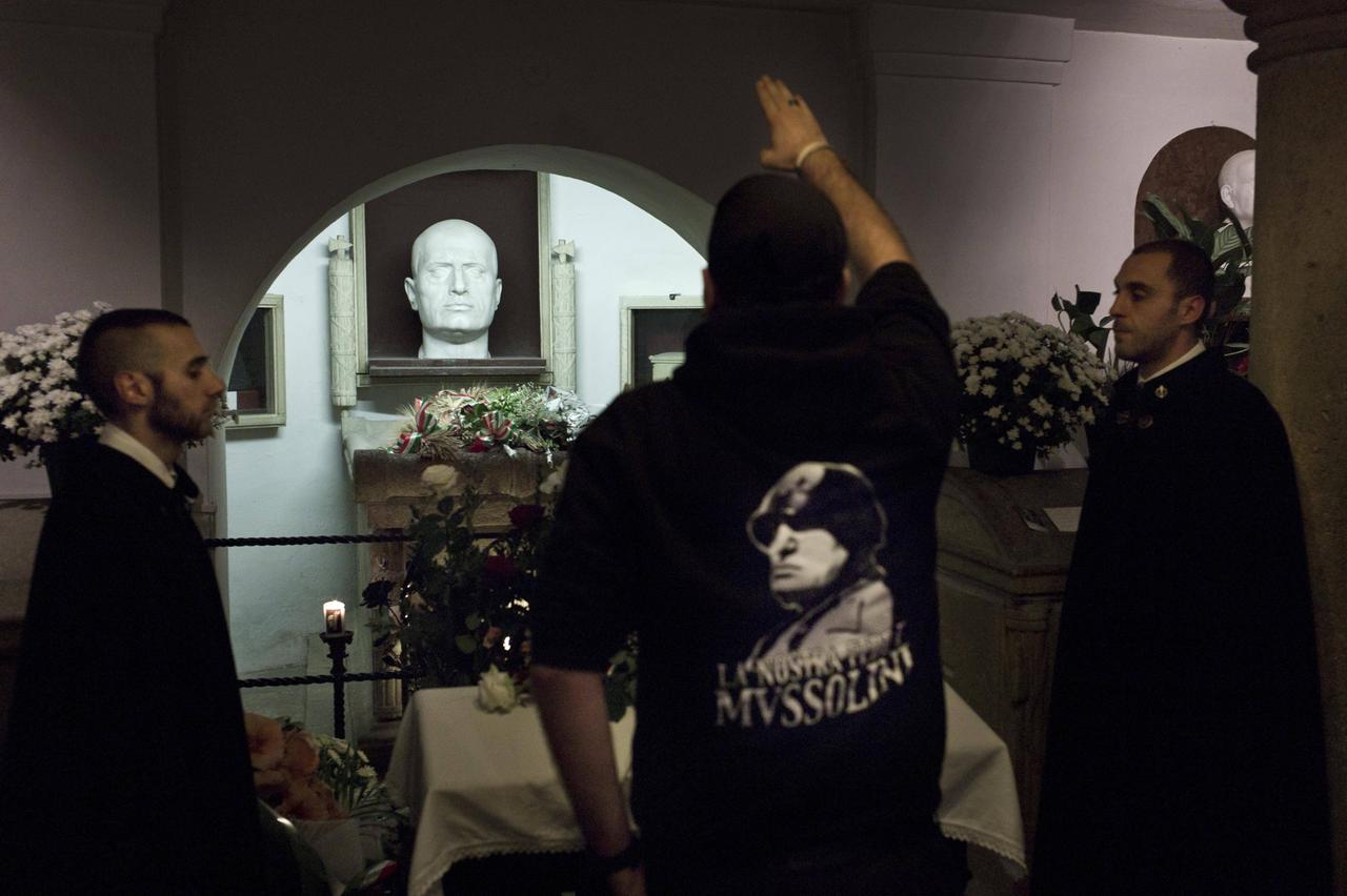 Ein Mann mit schwarzem Sweater, auf dessen Rückseite Mussolini abgebildet ist, streckt den gestreckten rechten Arm einer Mussolini-Büste an einer Gedenkstätte mit Blumen und Kerzen entgegen. 