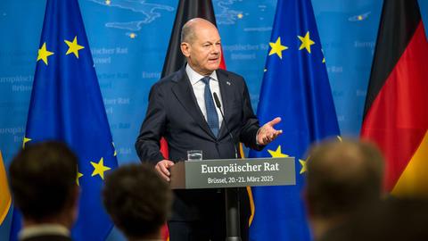 Olaf Scholz steht an einem Rednerpult mit der Aufschrift "Europäischer Rat - Brüssel, 20. - 21. März 2025". Im Hintergrund sieht man EU- und Deutschlandflagen
