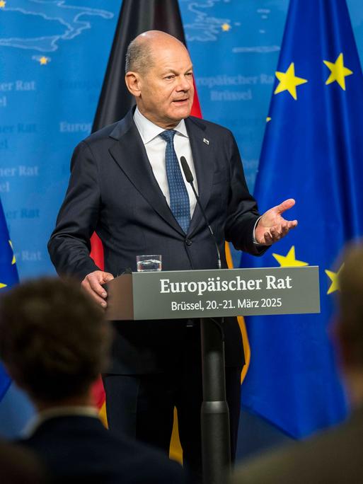 Olaf Scholz steht an einem Rednerpult mit der Aufschrift "Europäischer Rat - Brüssel, 20. - 21. März 2025". Im Hintergrund sieht man EU- und Deutschlandflagen