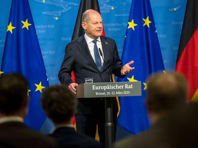 Olaf Scholz steht an einem Rednerpult mit der Aufschrift "Europäischer Rat - Brüssel, 20. - 21. März 2025". Im Hintergrund sieht man EU- und Deutschlandflagen
