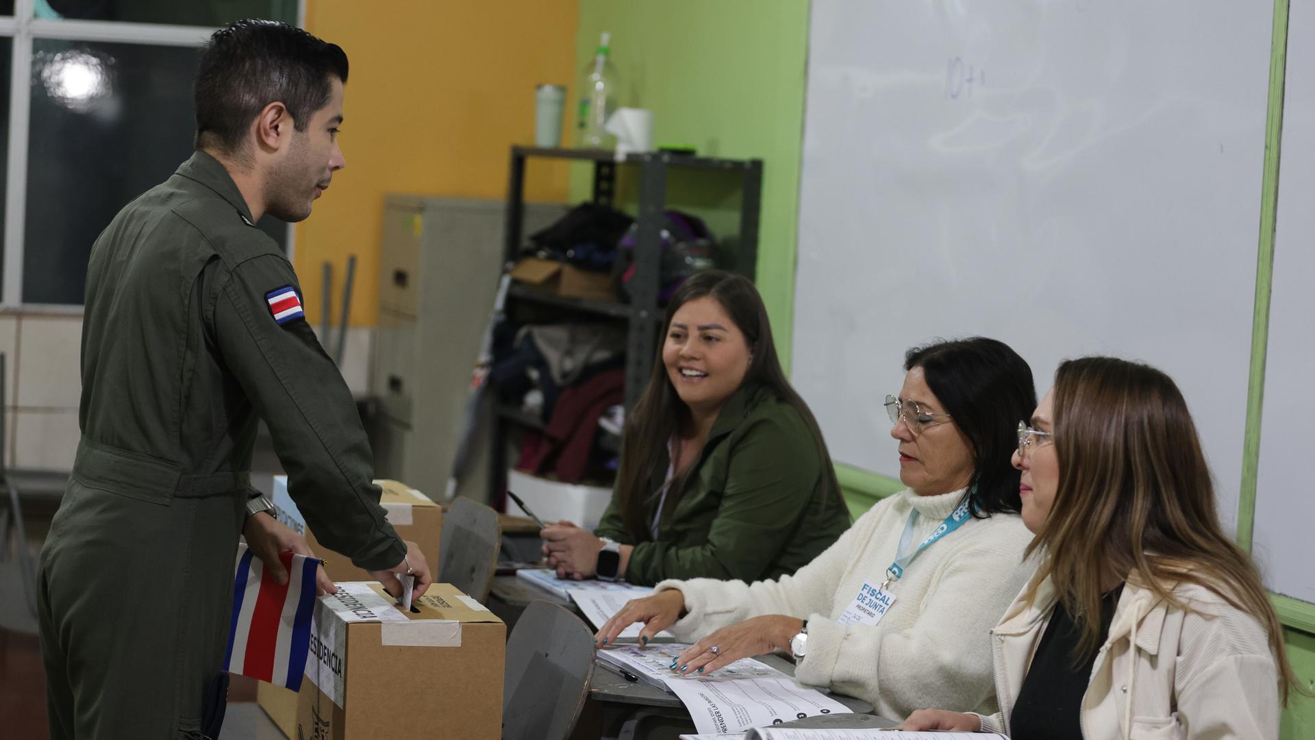Der Mann in Uniform steckt in einem Wahllokal den Stimmzettel in die Urne. An einem Tisch sitzen drei Wahlheferinnen. Der Mann in Uniform steckt in einem Wahllokal den Stimmzettel in die Urne. An einem Tisch sitzen drei Wahlheferinnen.