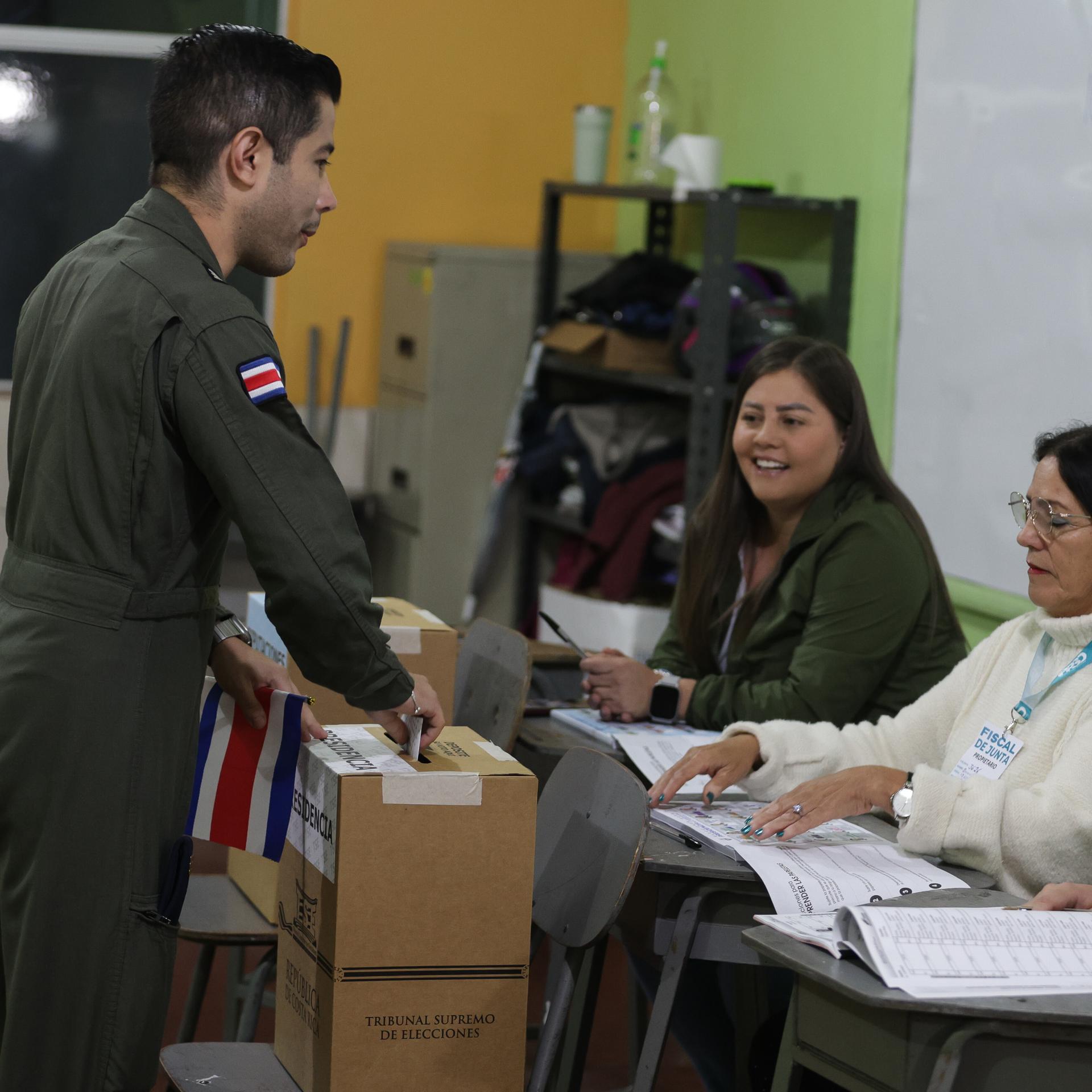 Der Mann in Uniform steckt in einem Wahllokal den Stimmzettel in die Urne. An einem Tisch sitzen drei Wahlheferinnen.