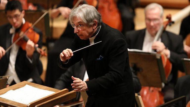 Ein Mann in einem Anzug hält einen Dirigentenstab, um ihn herum sitzen Musiker.