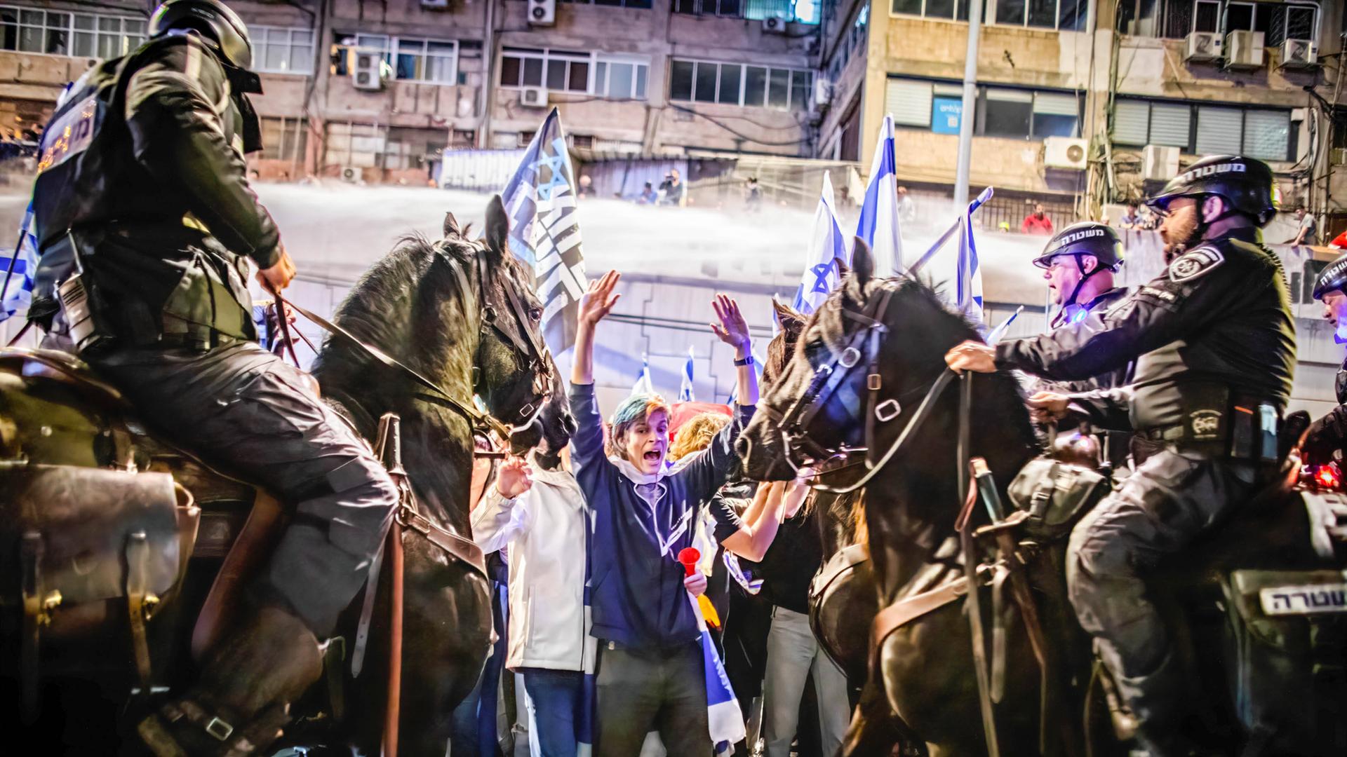 Das Foto zeigt einen israelischen Demonstranten mit erhobenen Händen. Polizisten auf Pferden bedrängen den Mann.