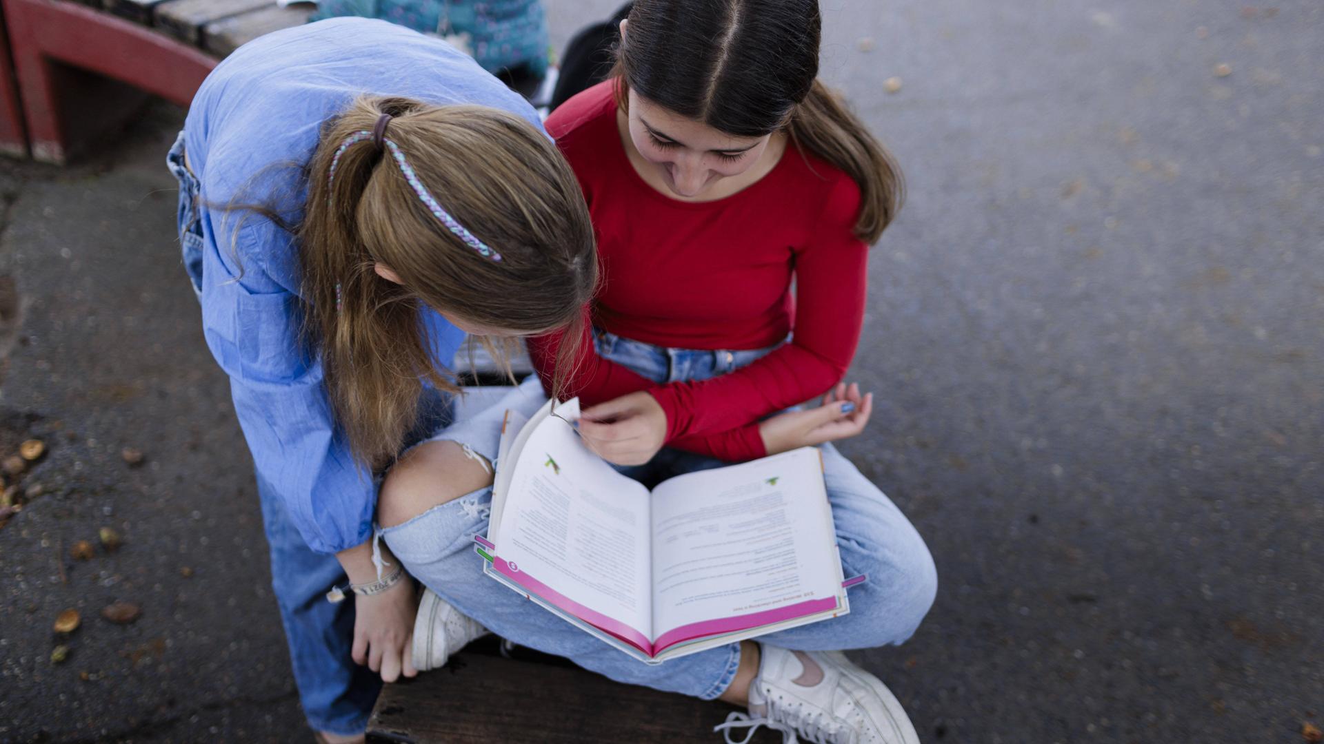 Zwei Mädchen sitzen über ein Buch gebeugt auf einem Schulhof in Deutschland.