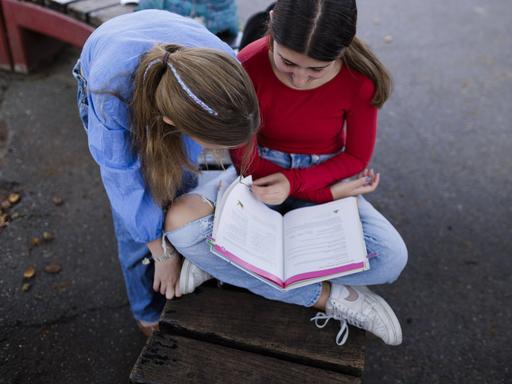 Zwei Mädchen sitzen über ein Buch gebeugt auf einem Schulhof in Deutschland.