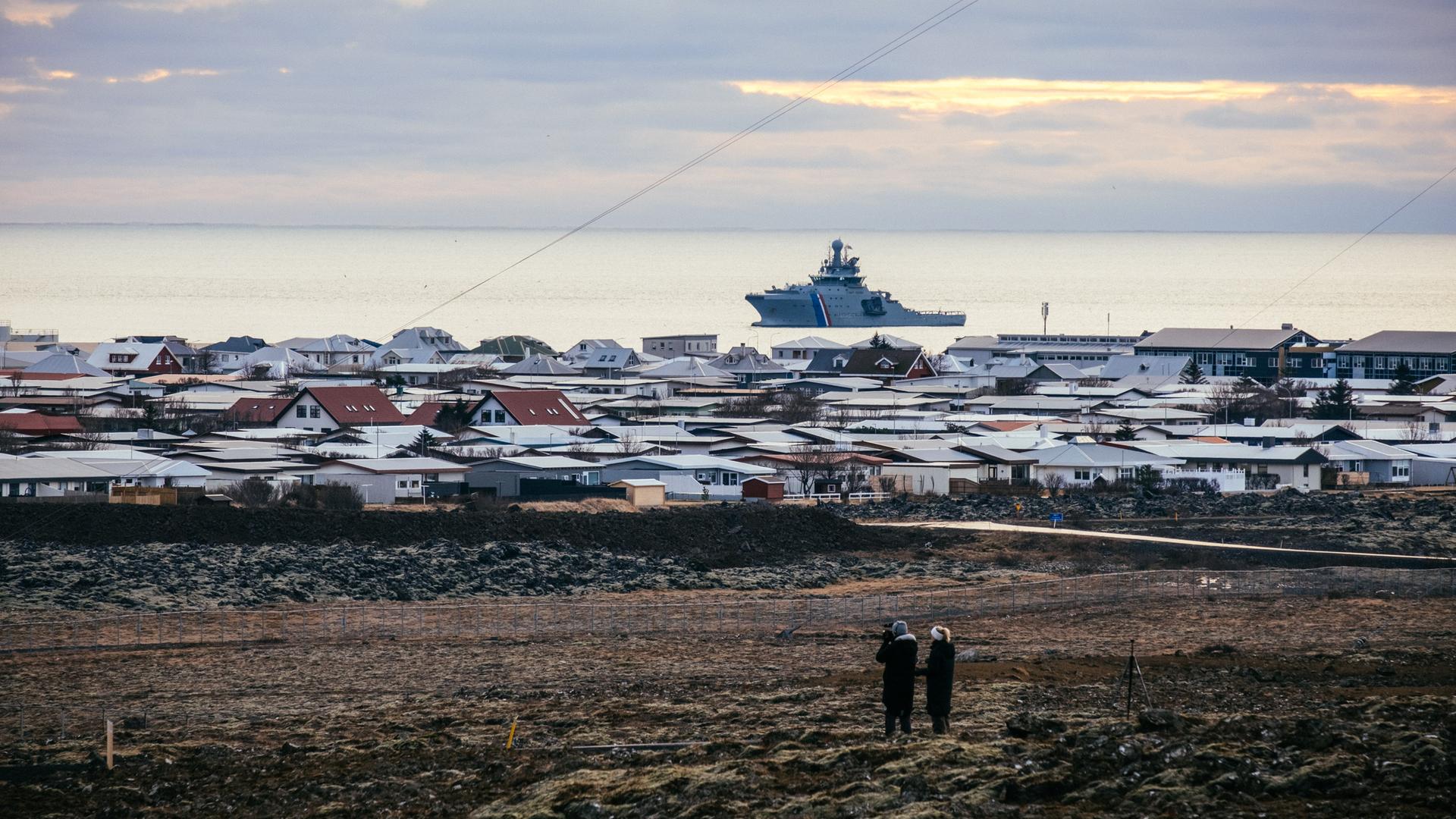 Ein Schiff der isländischen Küstenwache liegt vor Island im Meer. Auf dem Festland sind die Dächer von Häusern zu sehen. Ein Schiff der isländischen Küstenwache liegt vor Island im Meer. Auf dem Festland sind die Dächer von Häusern zu sehen.