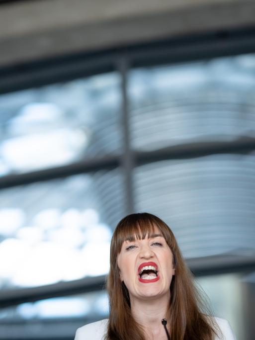 Heidi Reichinnek, Fraktionsvorsitzende Die Linke, gibt vor einem unscharfen Hintergrund aus Glasscheiben ein Statement ab. Ihr Kopf ist klein in der unteren Bildhälfte zu sehen. Sie scheint etwas zu rufen oder laut auszusprechen.
