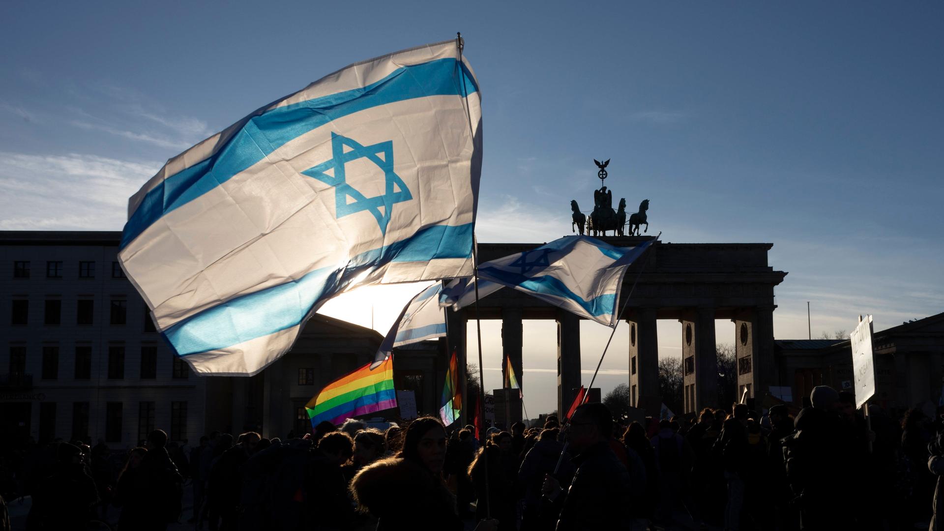 Eine israelische Fahne und eine Regenbogenfahne werden bei einer Demonstration geschwenkt. Im Hintergrund erkennt man das Brandenburger Tor in Berlin.
