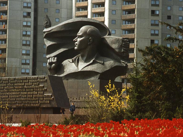 Das Ernst Thälmann Denkmal vor der Plattenbau-Kulisse in Berlin-Prenzlauer Berg. Im Vordergrund rote Blumen, im Hintergrund die Plattenbauten.