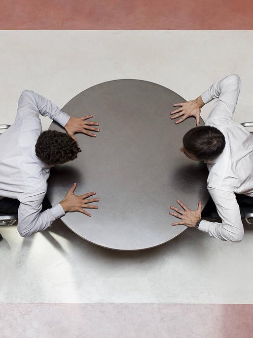 Aus der Vogelperspektive: Zwei Männer sitzen sich an einem Tisch gegenüber und streiten. 