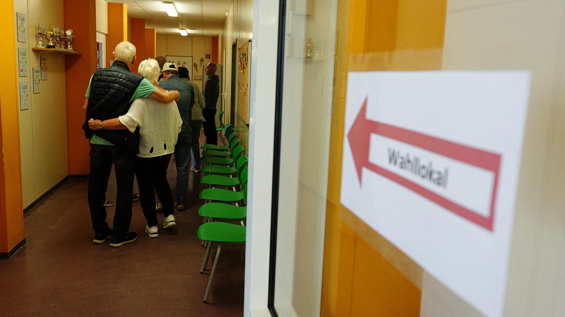 Mehrere Wählerinnen und Wähler warten in einem Wahllokal auf ihre Stimmabgabe, während ein Hinweisschild den Weg zum Wahlraum weist. Entlang des Flurs stehen Stühle. Mehrere Wählerinnen und Wähler warten in einem Wahllokal auf ihre Stimmabgabe, während ein Hinweisschild den Weg zum Wahlraum weist. Entlang des Flurs stehen Stühle.