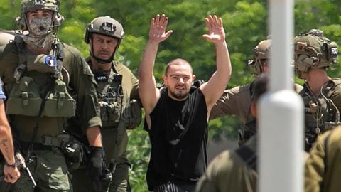 Ein Mann in ärmellosem, schwarzem Shirt streckt beide Arme in die Luft, er ist umringt von israelischen Soldaten, im Vordergrund steht ein Laternen- oder Fahnenmast