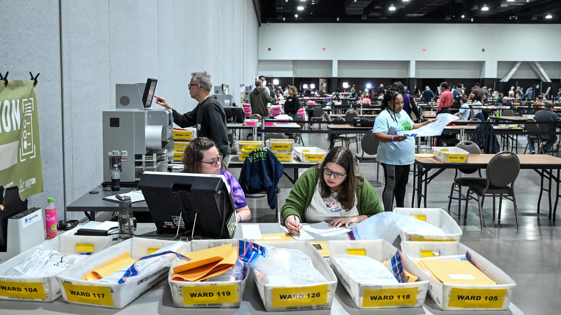 Eine Halle in Milwaukee, im Vordergrund zählen Wahlhelfer die Stimmen, im Hintergrund sitzen Menschen und geben ihre Stimme ab. Eine Halle in Milwaukee, im Vordergrund zählen Wahlhelfer die Stimmen, im Hintergrund sitzen Menschen und geben ihre Stimme ab.