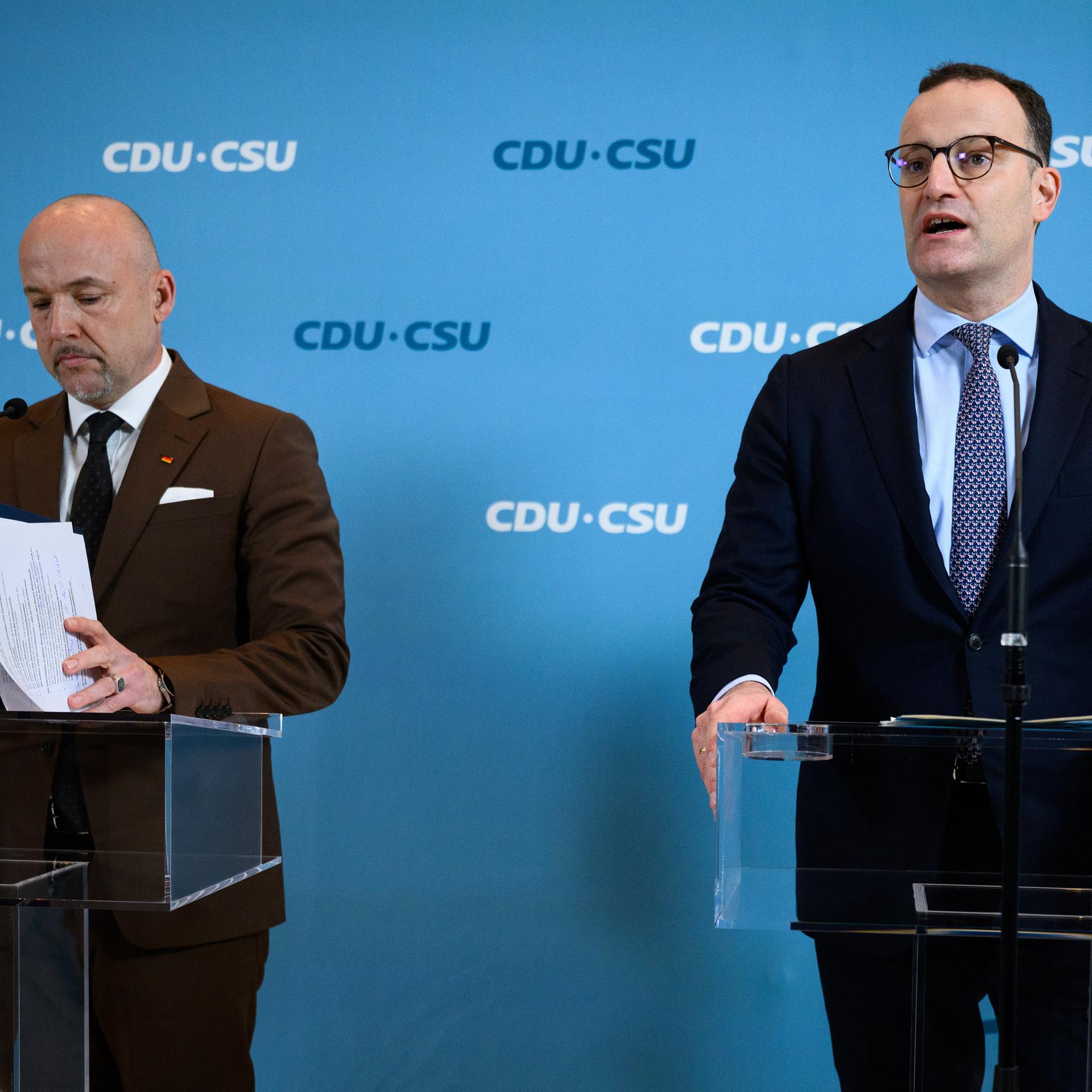 Jens Spahn und Alexander Hoffmann stehen an transparenten Redepulten und sprechen zu wartenden Journalisten. Im Hintergrund ist eine blaue Wand mit Logos der CDU/CSU-Bundestagsraktion zu sehen.