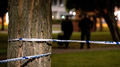 Polizeiabsperrband um einen Baum gewickelt, im Hintergrund zeichnen sich dunkel gekleidete Menschen ab.