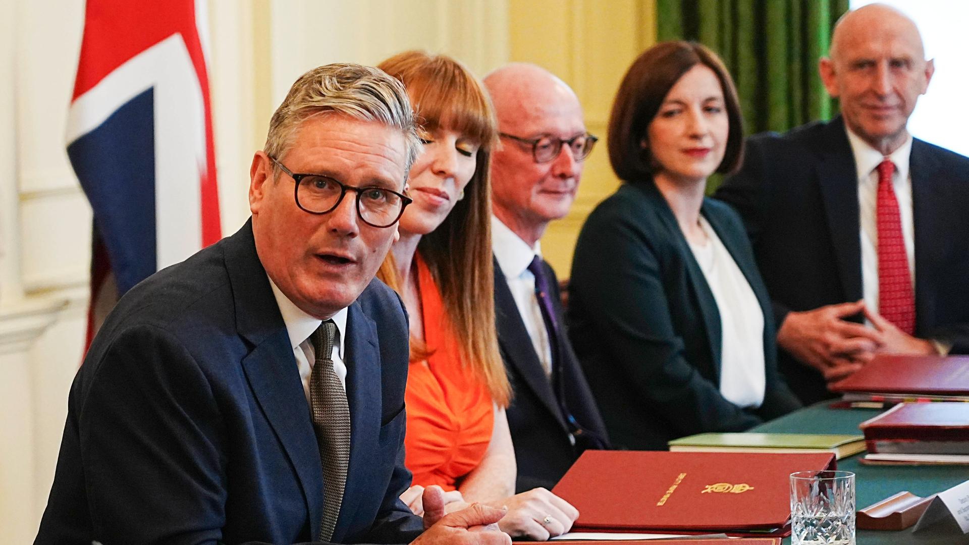 Premierminister Keir Starmer sitzt mit weiteren Regierungsmitgliedern an einem Tisch, vor ihnen liegen rote Mappen. Im Hintergrund ist eine britische Fahne zu sehen. 