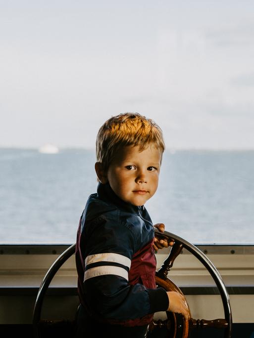Ein Junge am Steuerrad eines Schiffs. Durch die Scheibe sieht man den Horizont und das Meer.