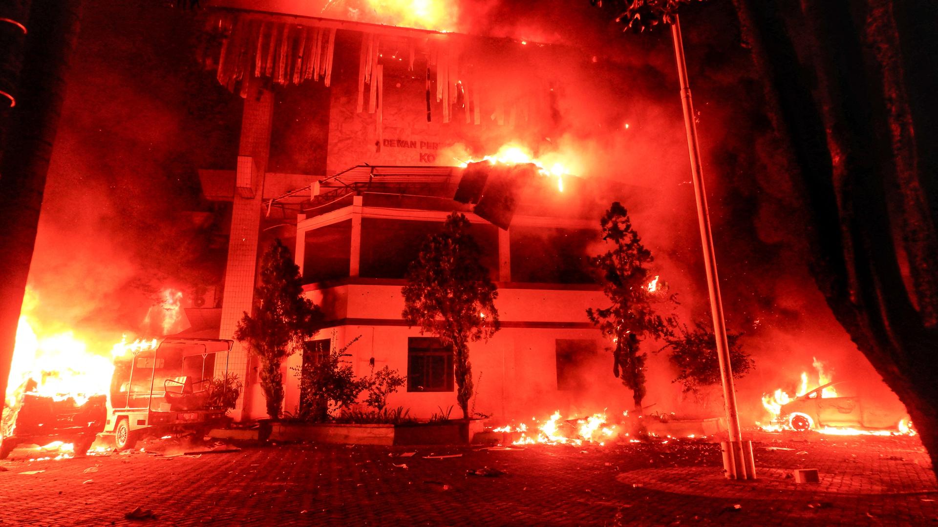 Das Foto zeigt ein mehrstöckiges Gebäude, das in Brand steht. Vor dem Gebäude stehen mehrere Fahrzeuge, die ebenfalls in Flammen stehen.