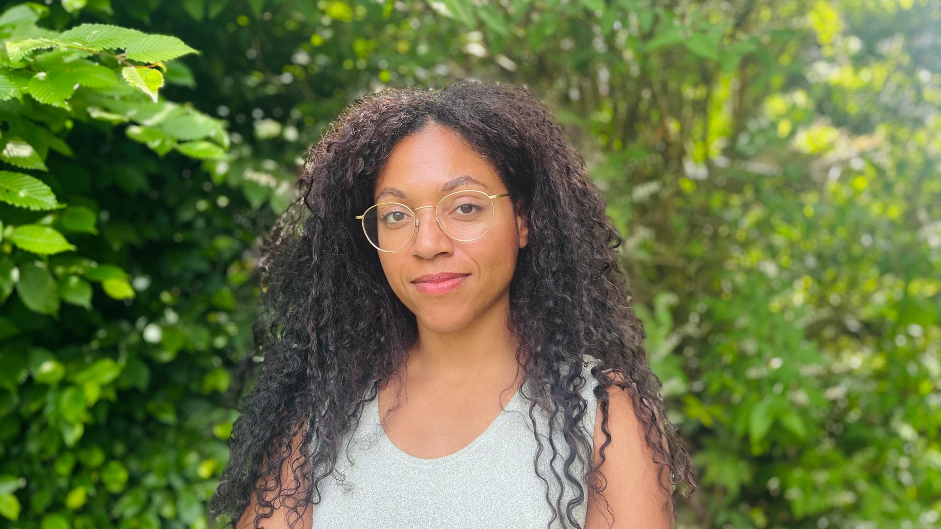 Esther trägt ihre langen Afrolocken offen. Sie hat eine goldene Brille auf und ein helles Top an. Um sie herum leuchtet das Grün der Baumblätter. Esther trägt ihre langen Afrolocken offen. Sie hat eine goldene Brille auf und ein helles Top an. Um sie herum leuchtet das Grün der Baumblätter.