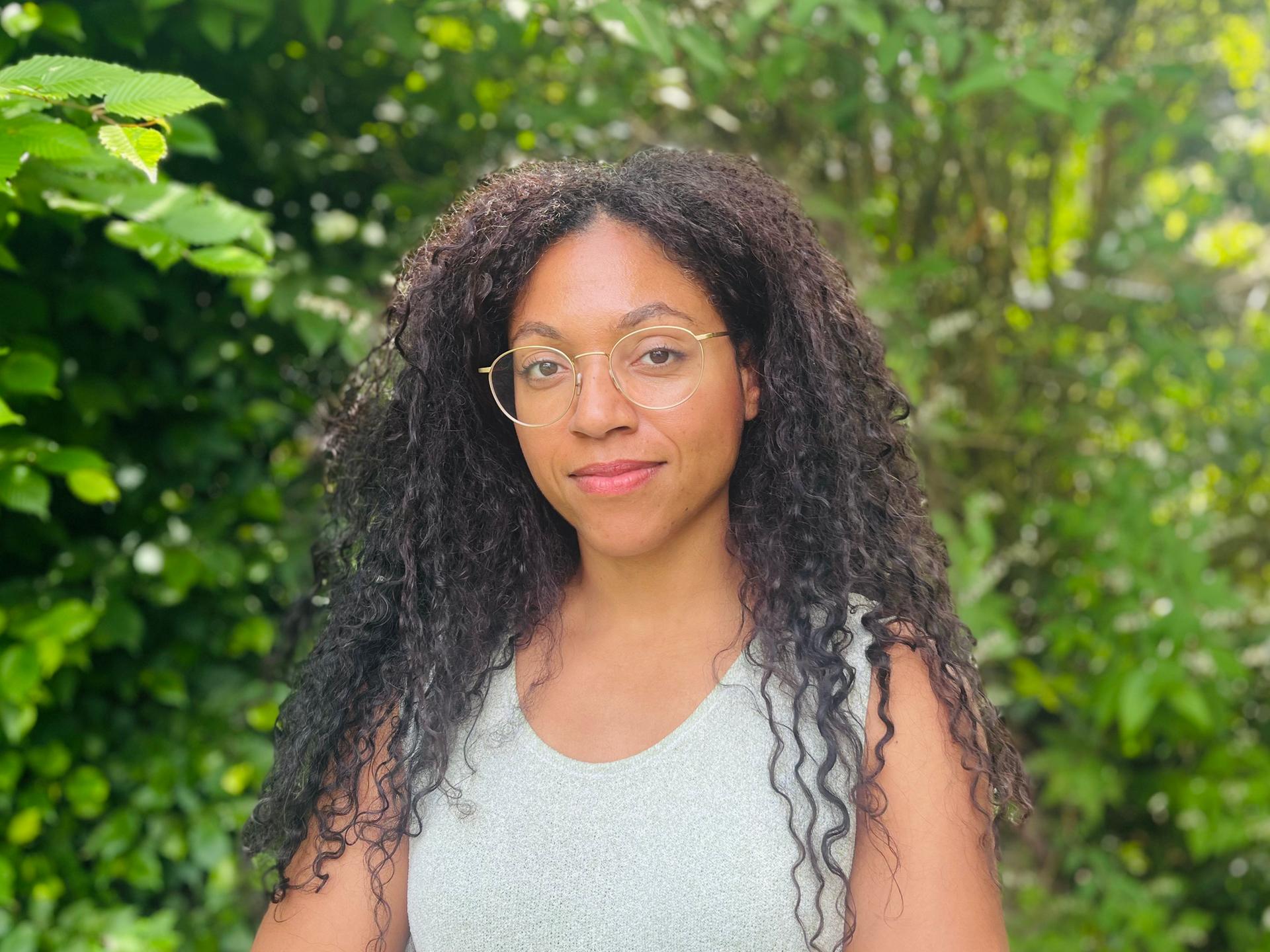 Esther trägt ihre langen Afrolocken offen. Sie hat eine goldene Brille auf und ein helles Top an. Um sie herum leuchtet das Grün der Baumblätter.