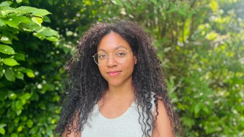 Esther trägt ihre langen Afrolocken offen. Sie hat eine goldene Brille auf und ein helles Top an. Um sie herum leuchtet das Grün der Baumblätter.