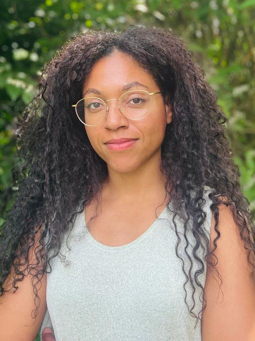 Esther trägt ihre langen Afrolocken offen. Sie hat eine goldene Brille auf und ein helles Top an. Um sie herum leuchtet das Grün der Baumblätter.