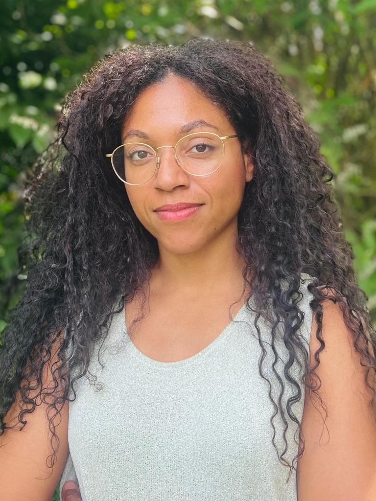 Esther trägt ihre langen Afrolocken offen. Sie hat eine goldene Brille auf und ein helles Top an. Um sie herum leuchtet das Grün der Baumblätter. Esther trägt ihre langen Afrolocken offen. Sie hat eine goldene Brille auf und ein helles Top an. Um sie herum leuchtet das Grün der Baumblätter.