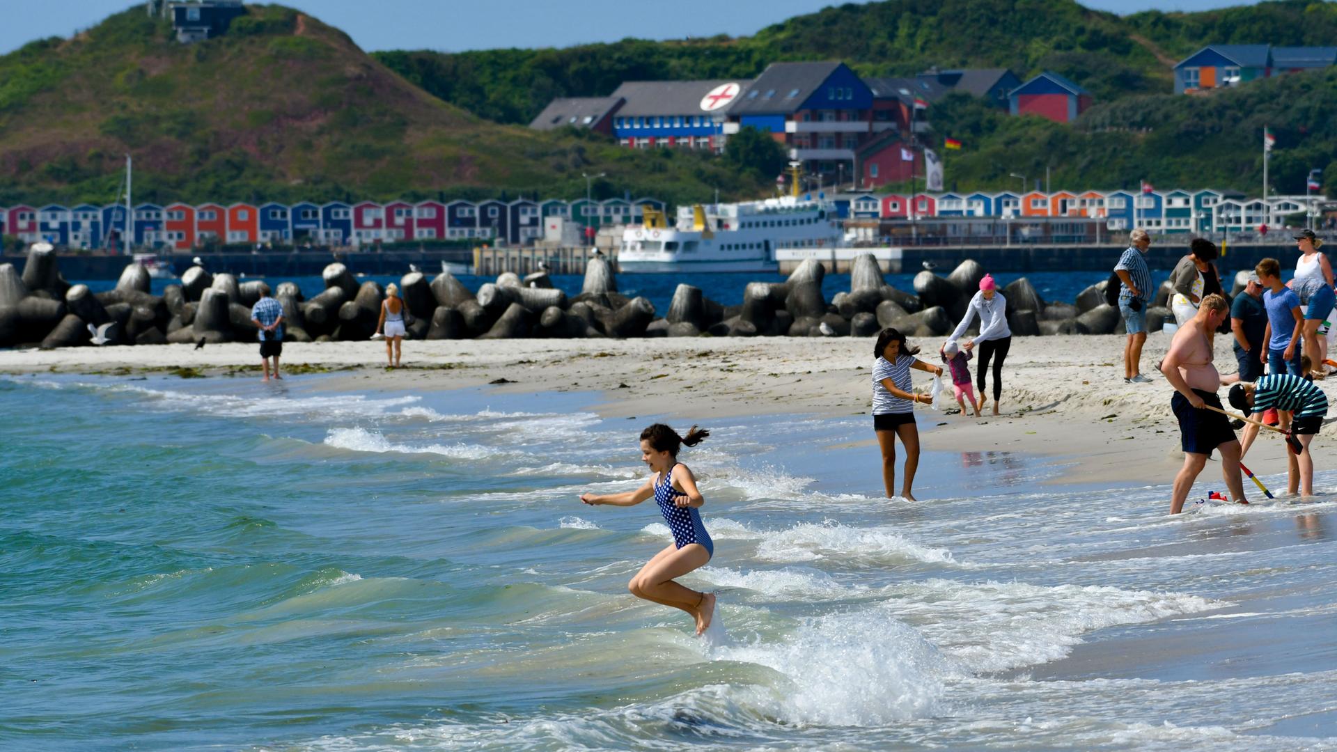 Menschen baden am Strand von Helogland im Meer. Im Hintergrund sieht man die berühmte Promenade mit den bunten Hummerbuden. 