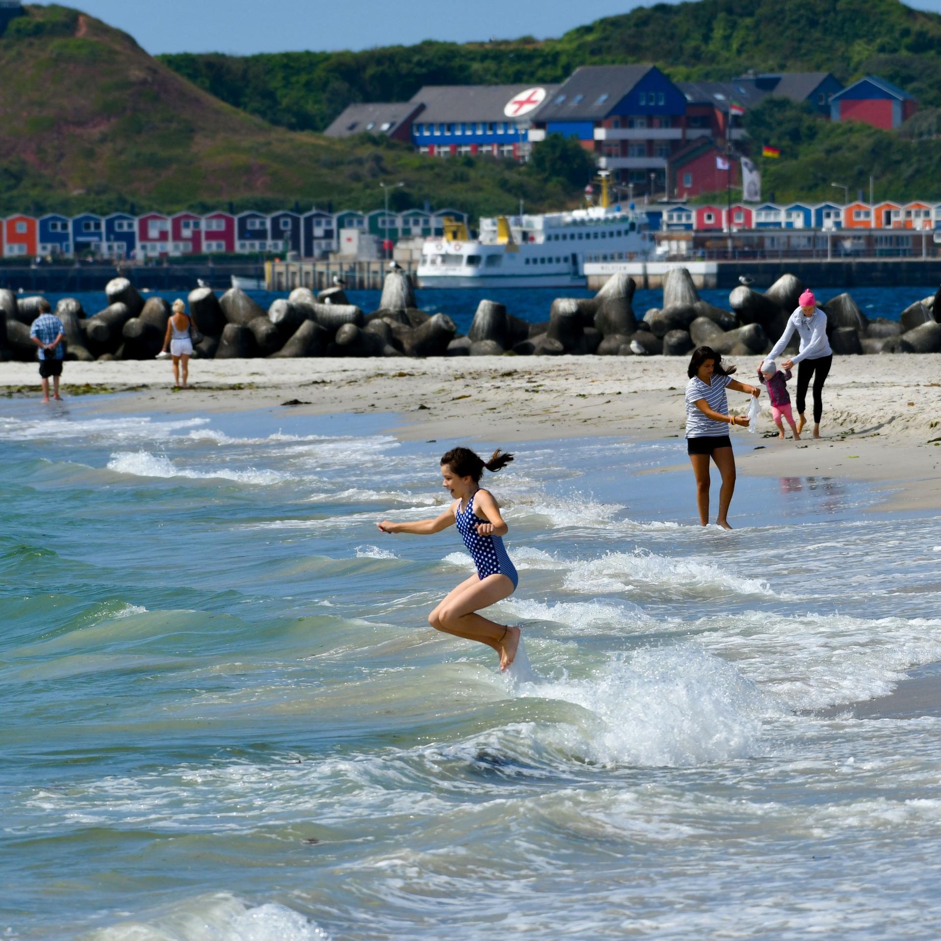 Menschen baden am Strand von Helogland im Meer. Im Hintergrund sieht man die berühmte Promenade mit den bunten Hummerbuden. 