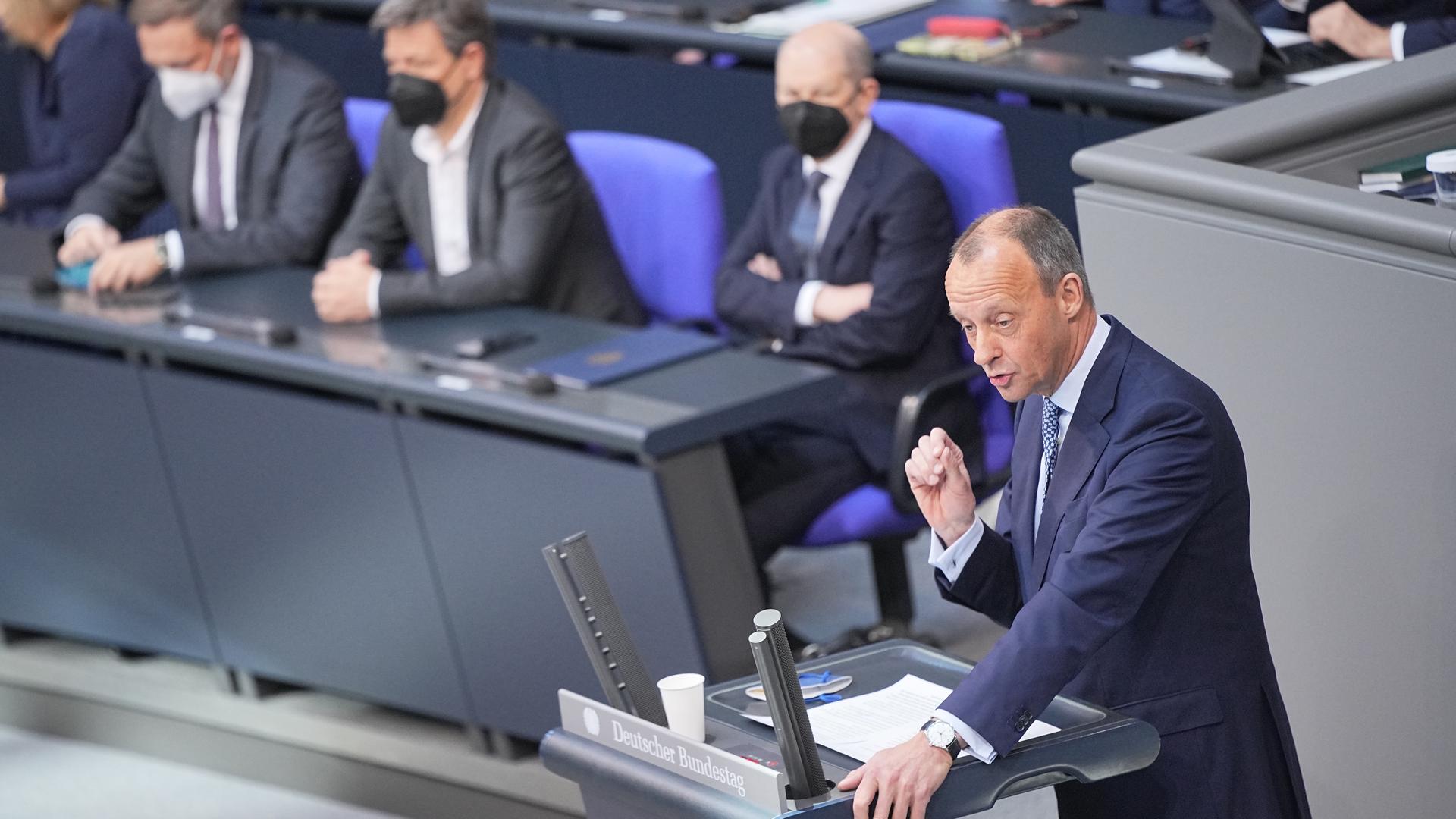 Im Bundestag steht Oppositionschef Friedrich Merz am Rednerpult, hinter ihm hören Kanzler Scholz und andere Mitglieder der Bundesregierung zu. Im Bundestag steht Oppositionschef Friedrich Merz am Rednerpult, hinter ihm hören Kanzler Scholz und andere Mitglieder der Bundesregierung zu.