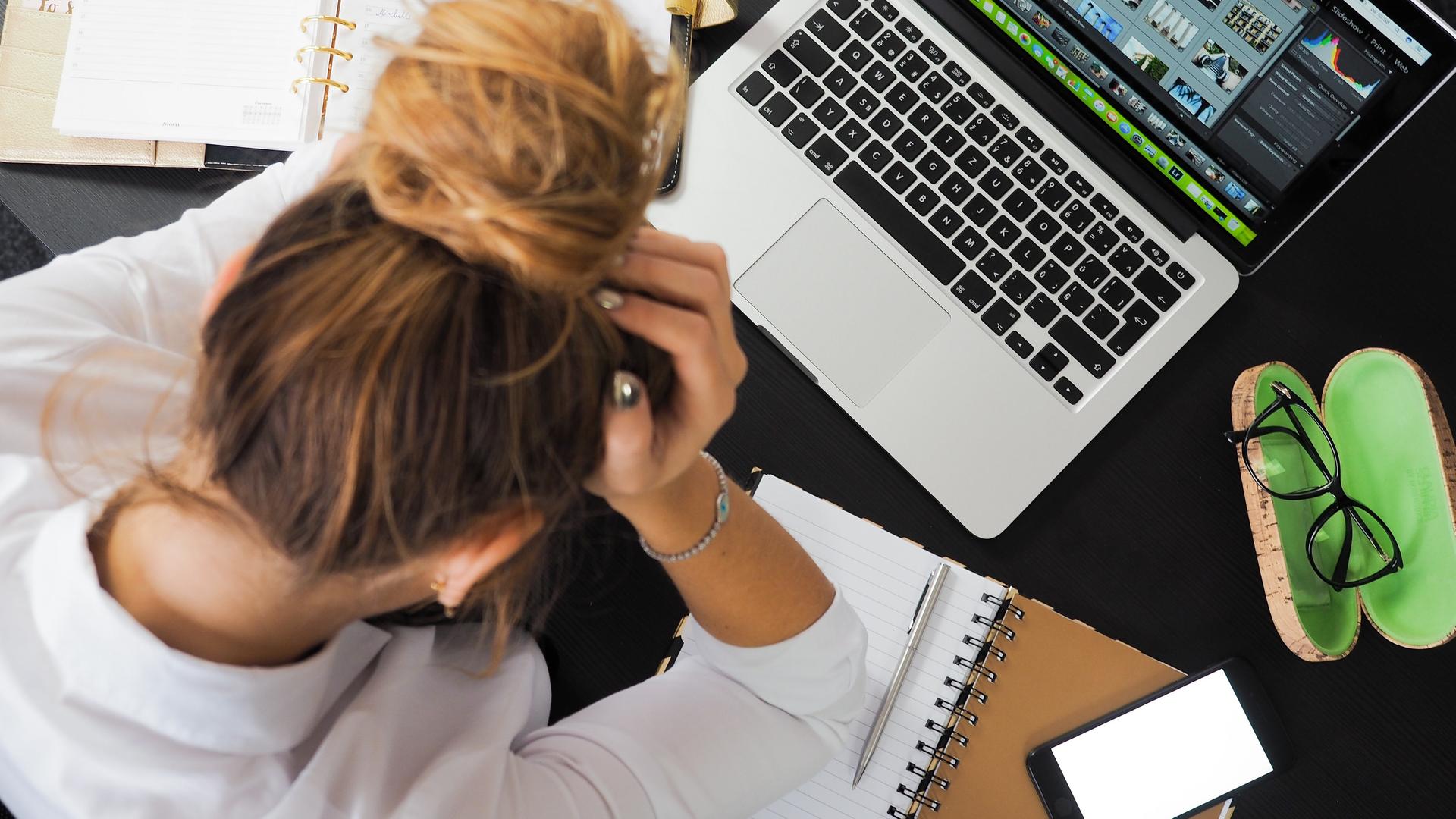 Eine Frau sitzt vor ihrem Laptop an einem vollen Schreibtisch und wirkt überarbeitet.