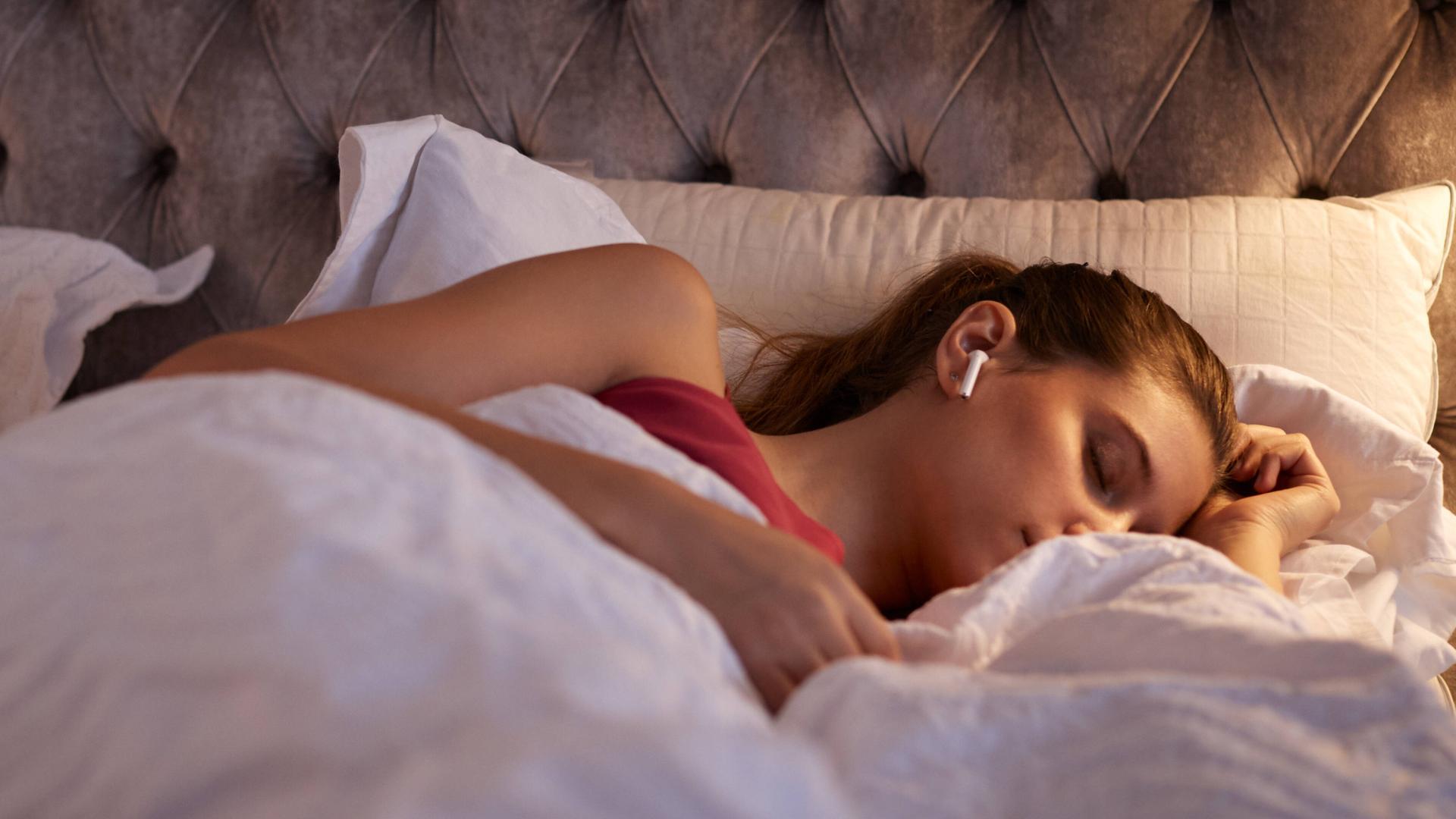 Eine junge Frau liegt mit geschlossenen Augen im Bett und hat sich eingekuschelt. In ihren Ohren hat sie einen weißen In-Ear-Kopfhörer.