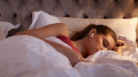 Eine junge Frau liegt mit geschlossenen Augen im Bett und hat sich eingekuschelt. In ihren Ohren hat sie einen weißen In-Ear-Kopfhörer.