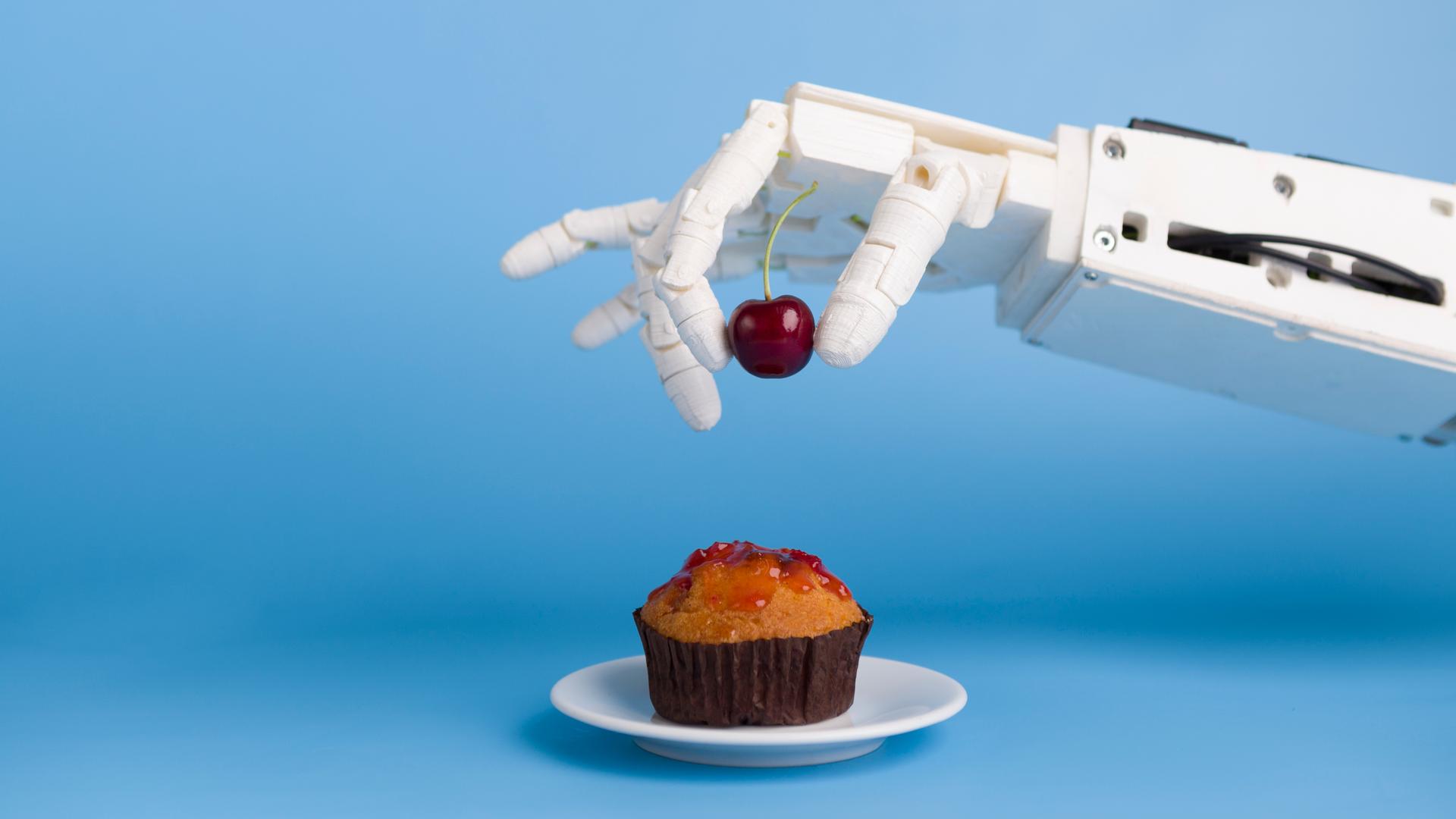 Eine Roboterhand setzt eine Kirsche auf einen Muffin. 