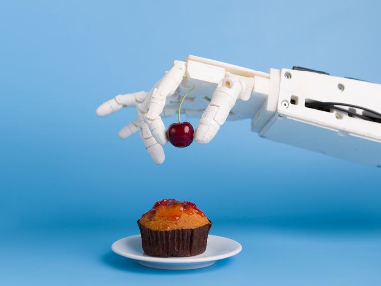 Eine Roboterhand setzt eine Kirsche auf einen Muffin. 