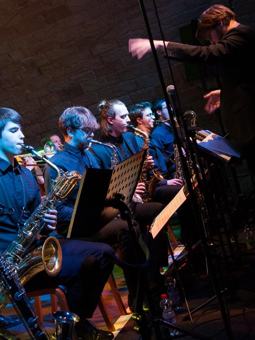 Eine Bigband mit mehreren Bläsern spielen sitzend an Notenpulten, davor der Dirigent