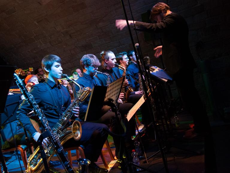 Eine Bigband mit mehreren Bläsern spielen sitzend an Notenpulten, davor der Dirigent